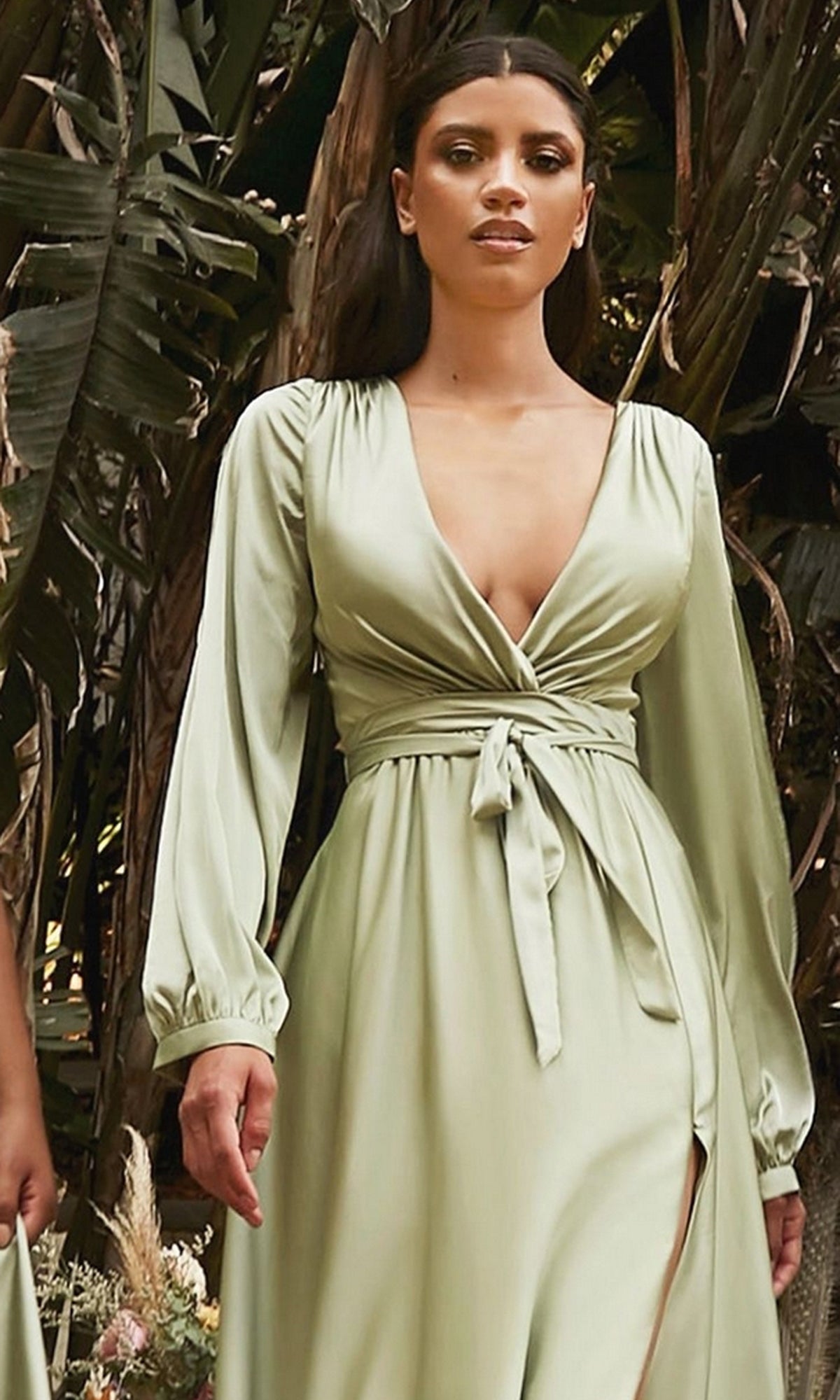 A woman wearing the Split Long Sleeve Long Formal Dress 7475 in light green, featuring a deep V-neckline, stands outdoors in front of large leafy plants.